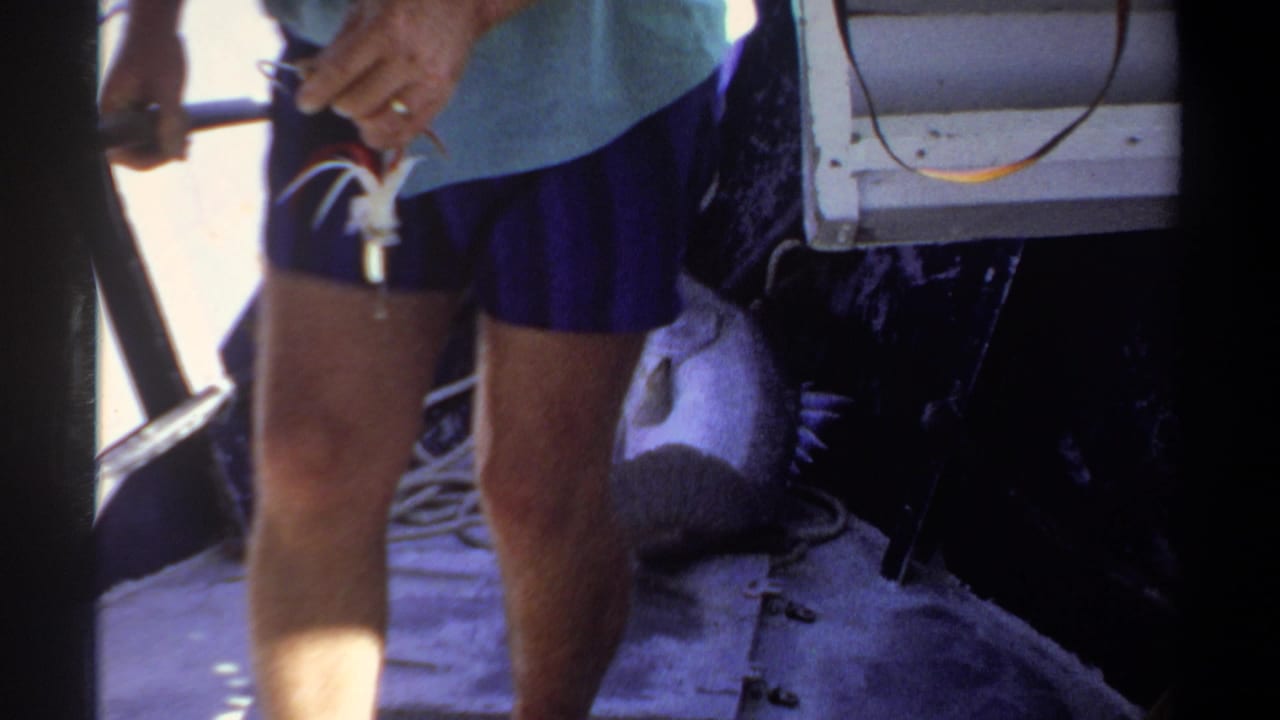 1969 Khartoum home-movie frame with the fisherman's legs, blue shorts, light shirt, and boat gear visible in close framing.