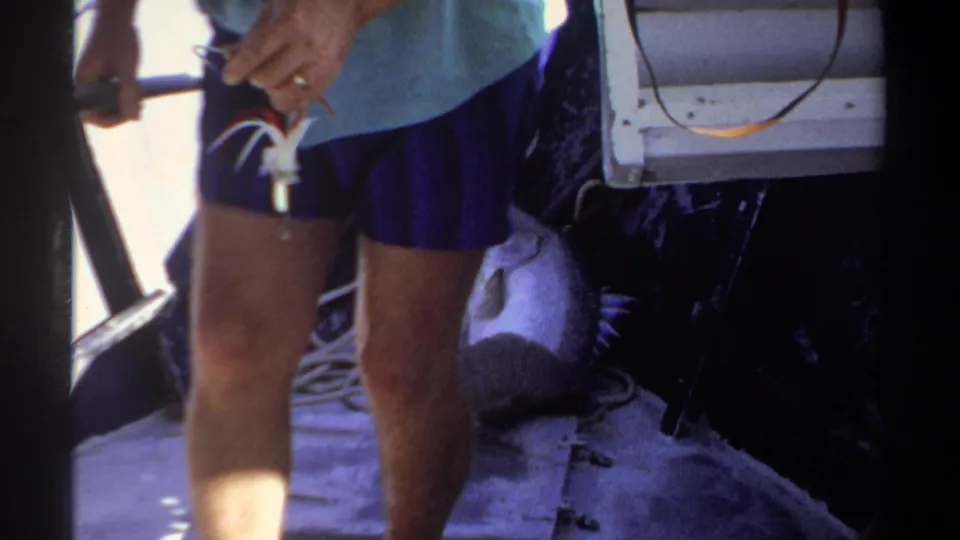 1969 Khartoum home-movie frame with the fisherman's legs, blue shorts, light shirt, and boat gear visible in close framing.