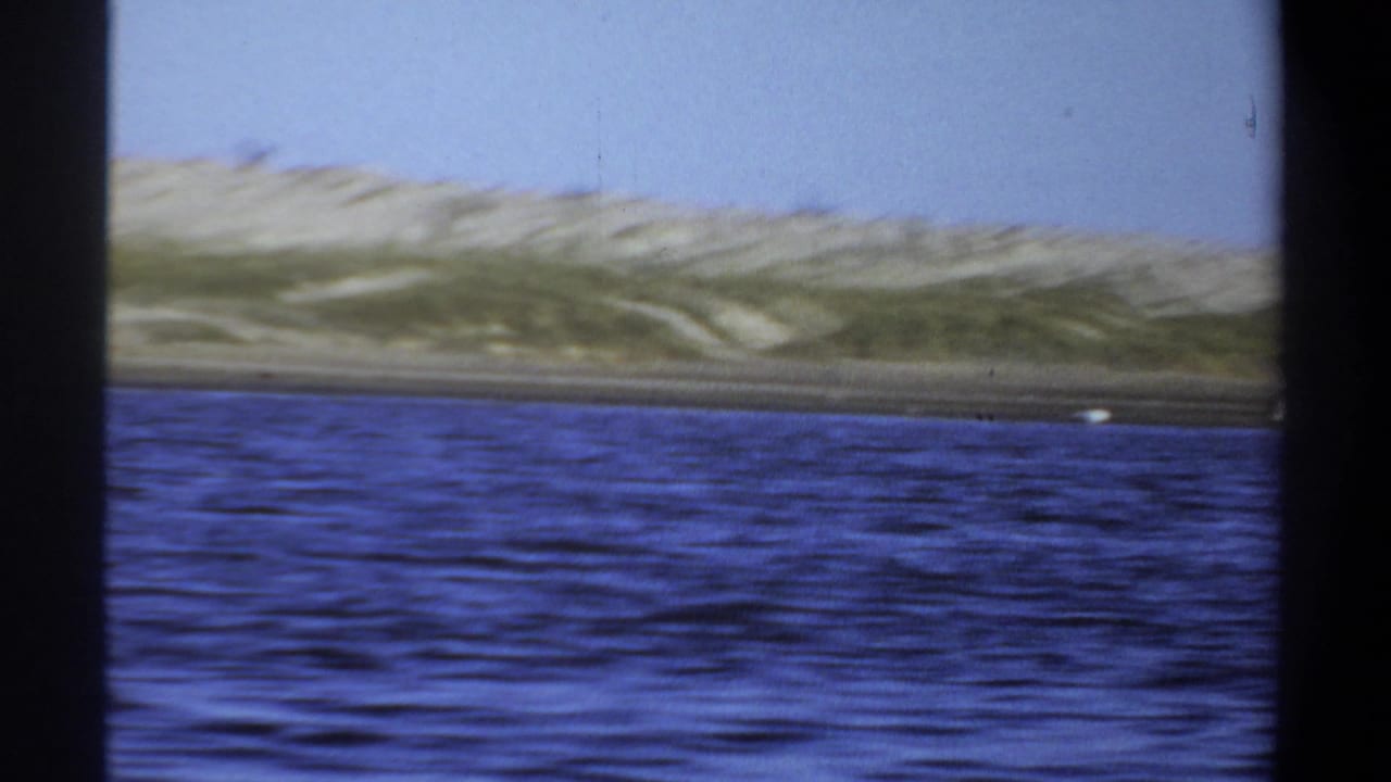 1969 Khartoum home-movie frame showing rippled blue water, sandy shoreline, sparse vegetation, and distant flamingos.