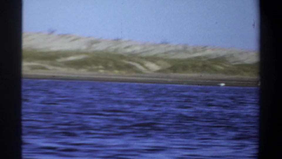 1969 Khartoum home-movie frame showing rippled blue water, sandy shoreline, sparse vegetation, and distant flamingos.