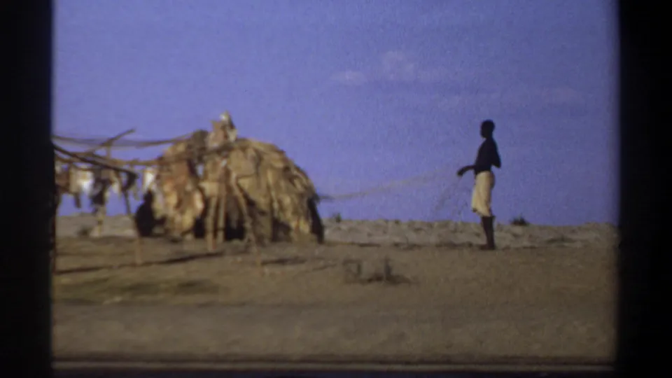 1969 Khartoum footage frame with a man standing near a thatched hut and wooden drying rack on a sparse riverside bank.