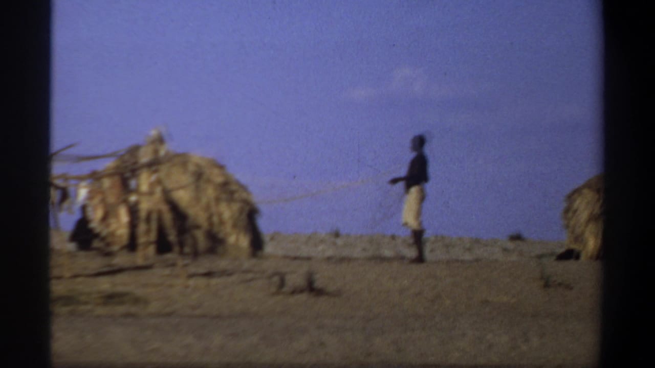 1969 Khartoum-area home-movie frame showing a man walking past straw-covered huts on dry open ground.