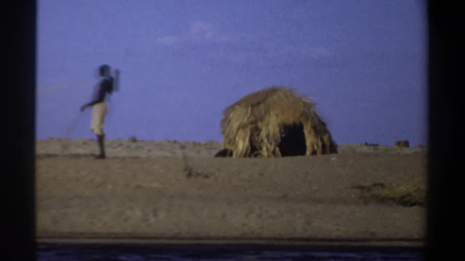 Distant figure stands or walks near a straw-covered hut in 1969 Khartoum home-movie footage.