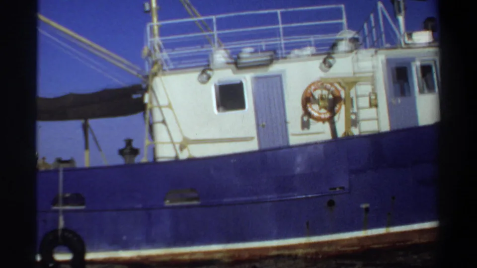 1969 Sudan 8mm frame of a blue-and-white boat cabin with deck railings, a lifebuoy, and rigging against clear sky.