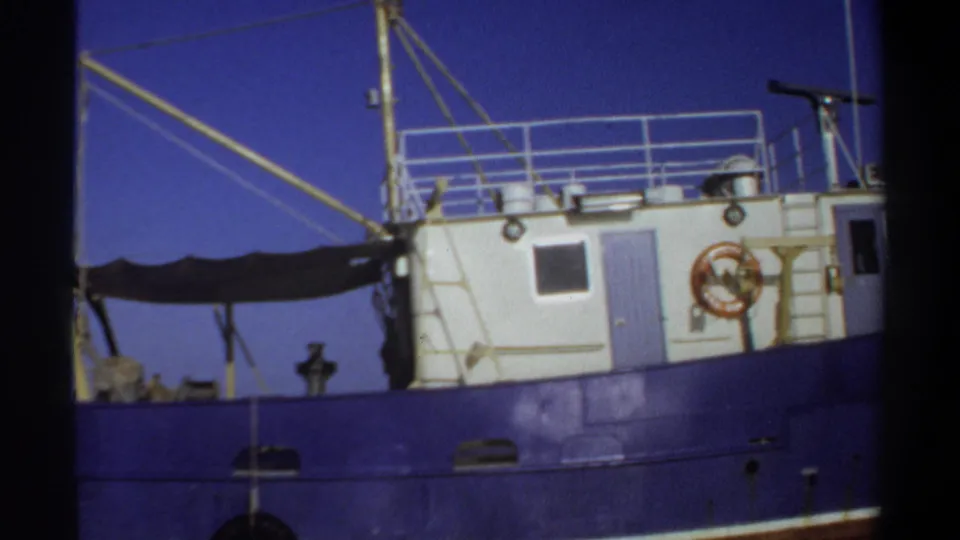 1969 Sudan 8mm frame showing the side of a blue-and-white boat, including cabin windows, a blue door, lifebuoy, and railings.