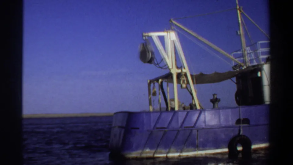 1969 Sudan-listed home-movie frame of a blue vessel on open water, showing yellow lifting gear, rigging, fenders, and a low distant shoreline.
