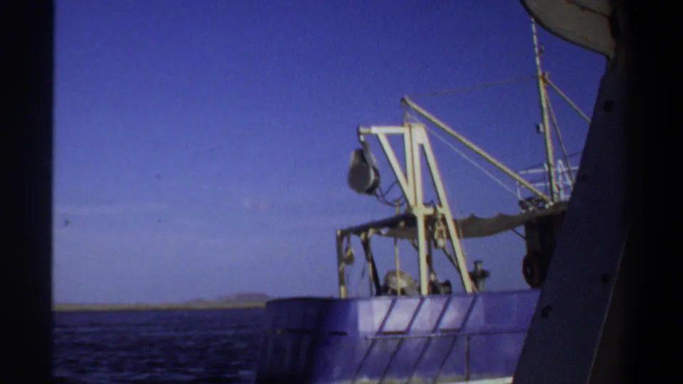 1969 Sudan-listed 8mm frame with a nearby boat's structure partly blocking the view of a blue vessel, yellow crane, rigging, water, and shoreline.