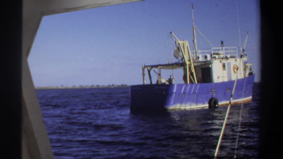 1969 Sudan 8mm frame of a blue boat with a white cabin and rigging, viewed across rippled water from another vessel.