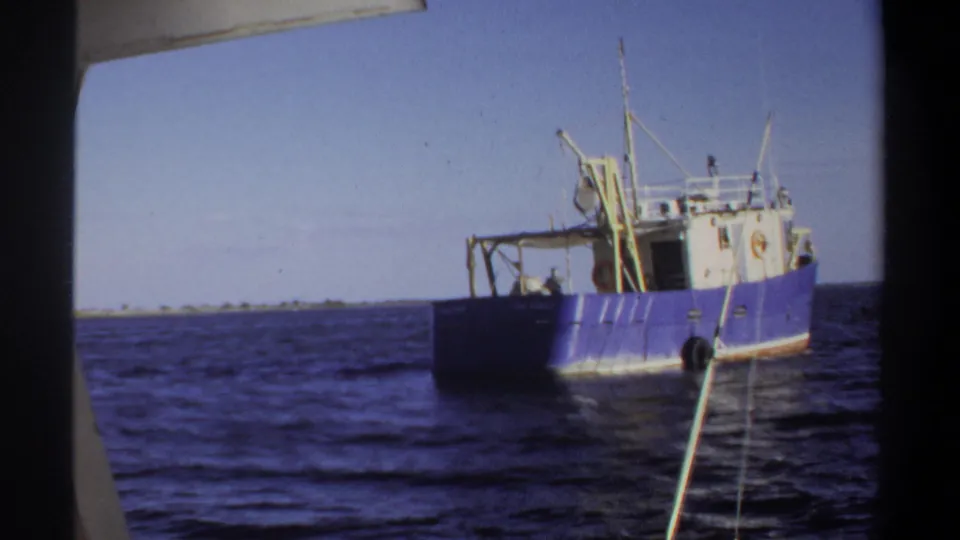 1969 Sudan 8mm frame showing the blue and white vessel more centered, with deck equipment, rigging, and nearby boat edges visible.