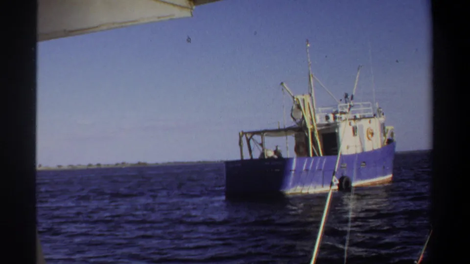 1969 Sudan 8mm frame with the blue and white boat at right, open water across the foreground, and a low shoreline in the distance.