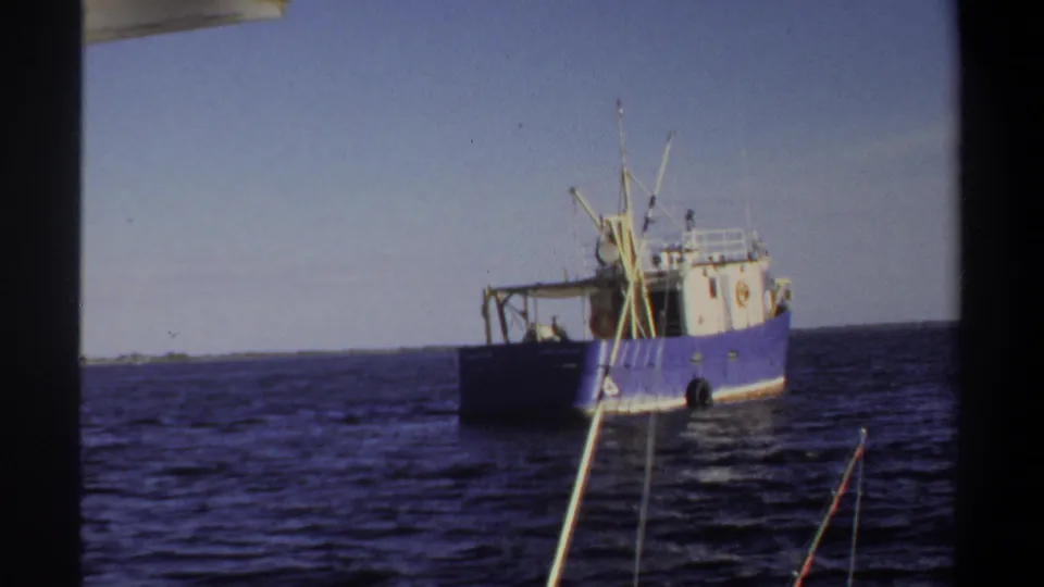 The blue and white fishing boat remains in view across calm water, with rods and a distant low shore visible.