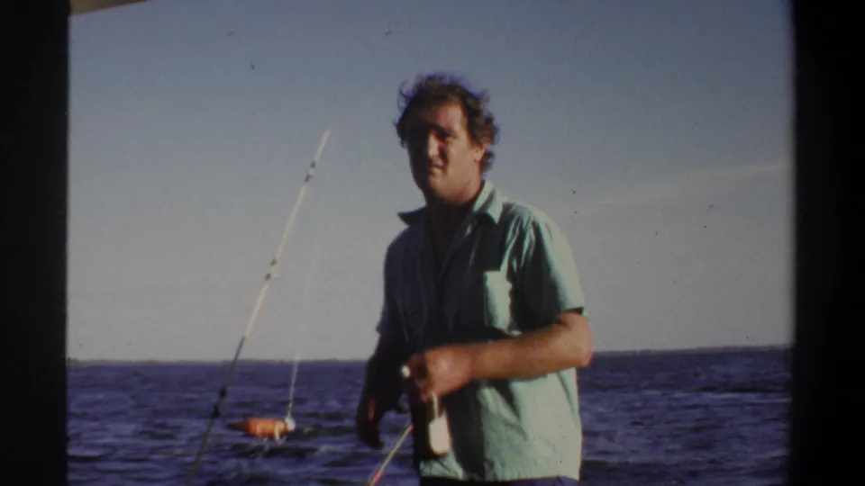Adult man in a green shirt stands on the deck during a 1969 Sudan fishing outing, with open water and rods behind him.