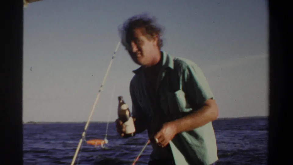 Adult man on a boat holds a bottle toward the camera, with fishing rods and the open sea behind him.