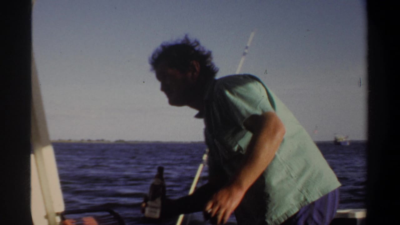 Man in a green shirt leans on a boat while holding a bottle near open water.