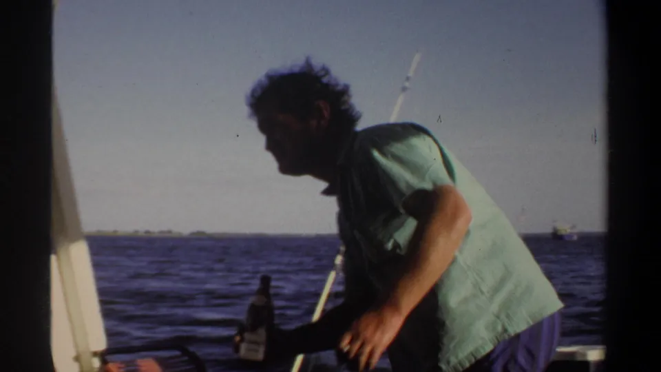 In 1969 Khartoum footage, a man in a green shirt leans forward on a boat with a bottle, with water and distant shore behind him.