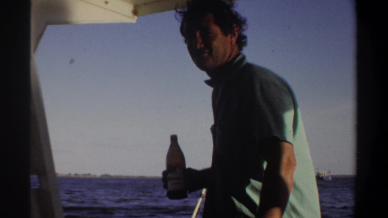 Man stands under a boat canopy holding a bottle with open water behind him.