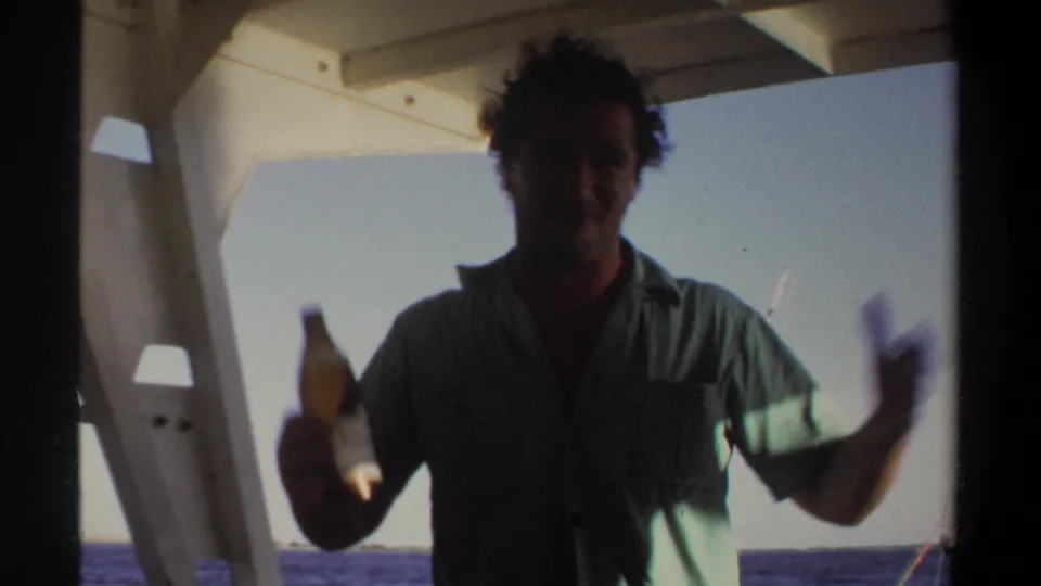 In the 1969 Sudan home movie, a backlit man holds a bottle and gestures beneath a boat canopy on open water.
