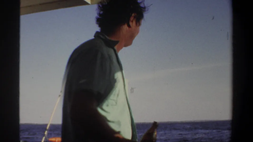 A man in a green shirt turns toward the water on a boat in Khartoum, with a fishing rod and distant shoreline visible.