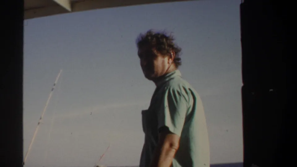 Adult man in a light green shirt looks partly toward the camera on a boat, with fishing rods and open water in the background.