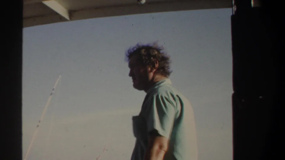 Profile view of an adult man standing on a boat in wind, with fishing rods and open water visible in the frame.