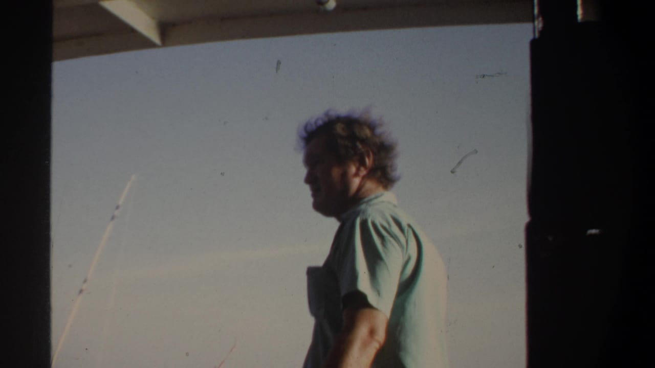 In 1969 Khartoum home-movie footage, a man in a pale green shirt stands on a boat with fishing rods visible against the sky.