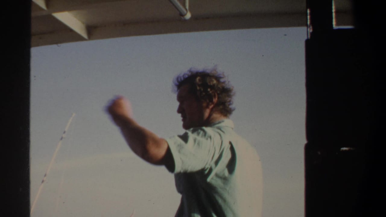 Man on a boat raises one arm while standing near fishing rods against the sky.