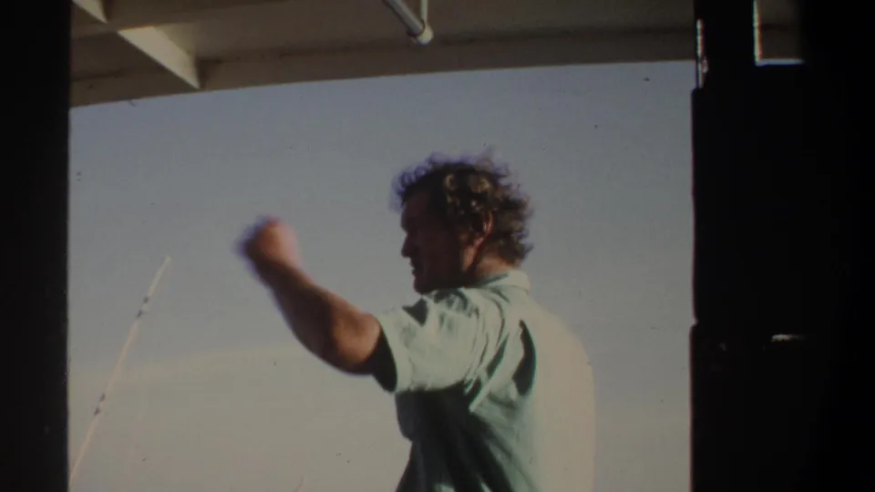 The man on the boat gestures with one arm near fishing rods in 1969 Khartoum home-movie footage.