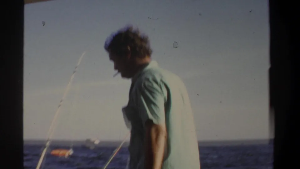 A man with a cigarette stands near a fishing rod on a boat in 1969 Khartoum footage, with open water in the background.