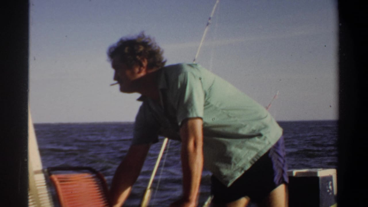 1969 home-movie frame of an adult man on a small boat, leaning forward with a cigarette as open water fills the background.