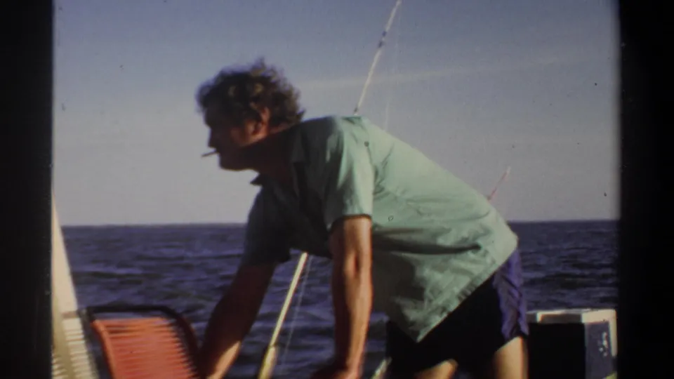 1969 home-movie frame of an adult man on a small boat, leaning forward with a cigarette as open water fills the background.