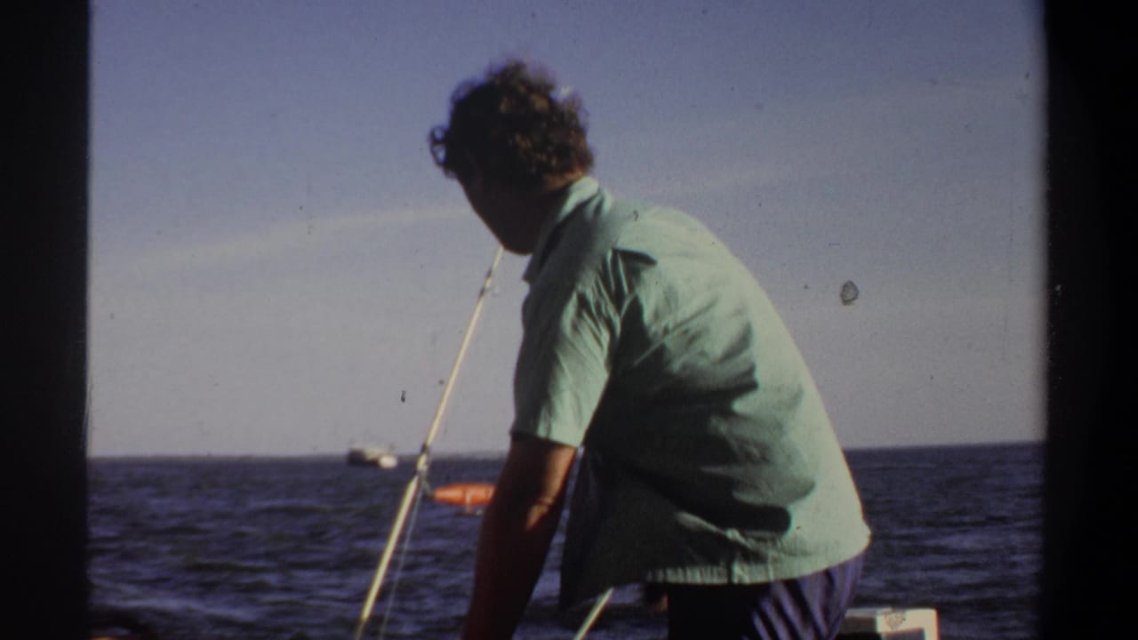 Man seen from behind on a boat near a fishing rod, with open water and sky beyond.