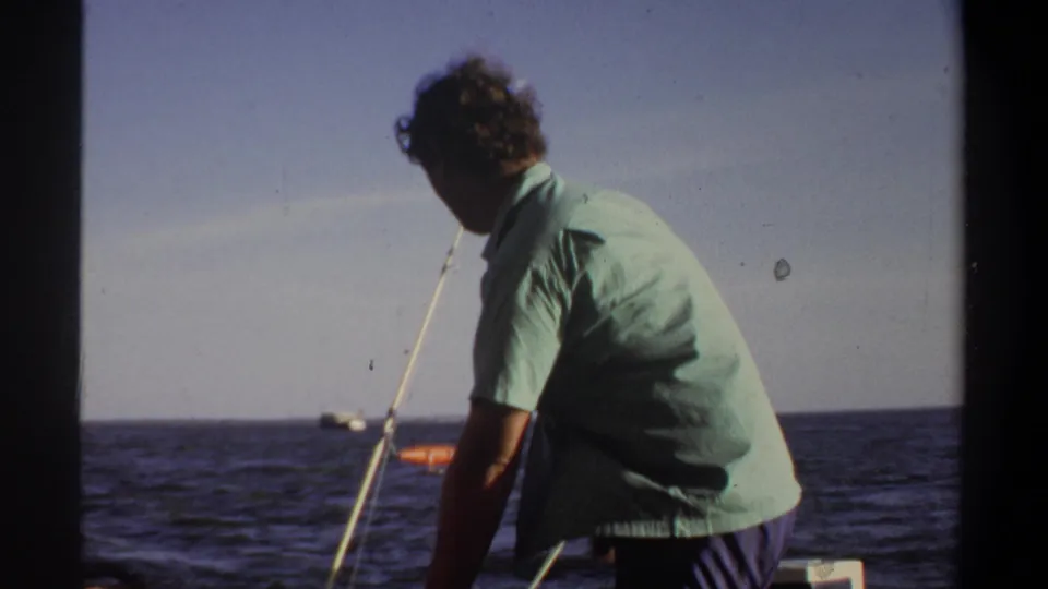 Adult man in a light green short-sleeved shirt stands on a boat beside a fishing rod, facing open water in the 1969 reel.