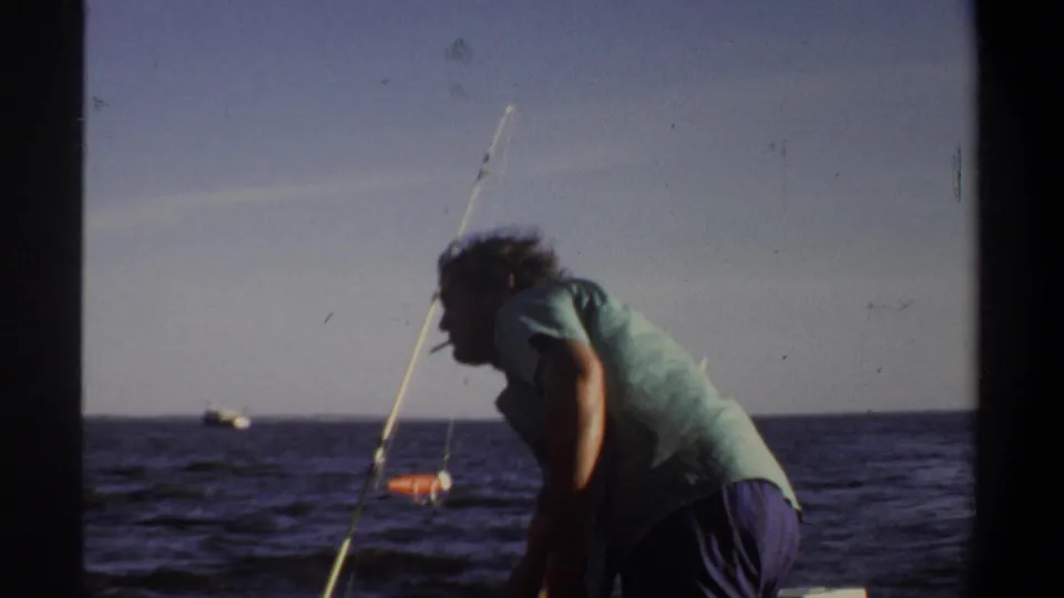 Adult man on a small boat leans near a fishing rod while a distant boat-like shape sits on the water under a clear sky.