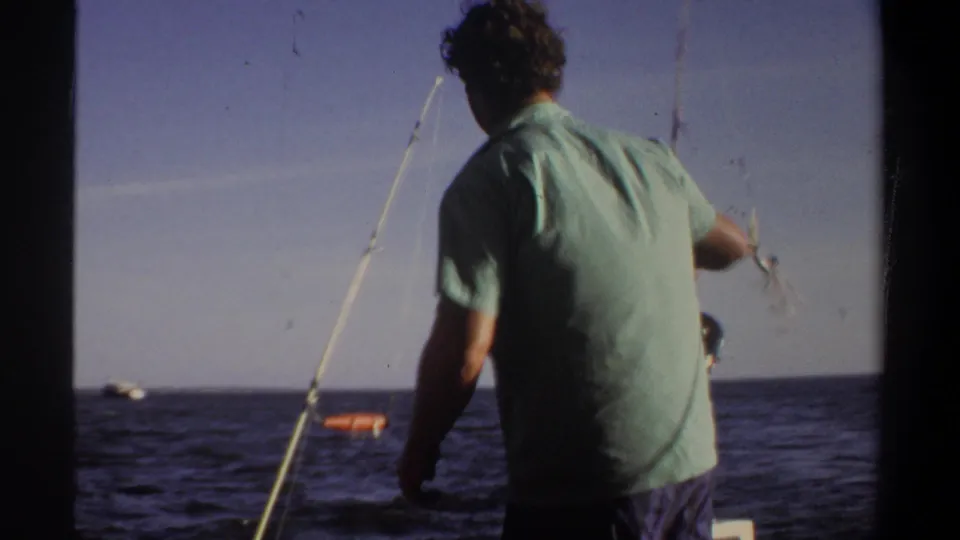 1969 home-movie frame showing an adult man from behind on a boat, with fishing rods angled toward open water and a clear sky.