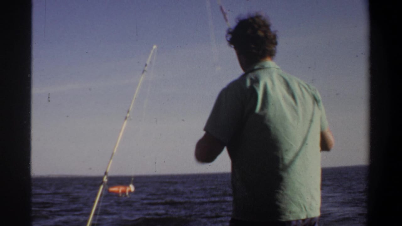 1969 Sudan-associated 8mm frame showing a man in a light green shirt handling a fishing rod near open water.