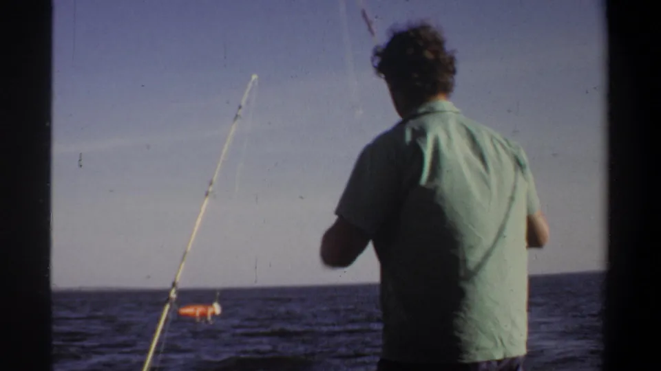 1969 Sudan-associated 8mm frame showing a man in a light green shirt handling a fishing rod near open water.