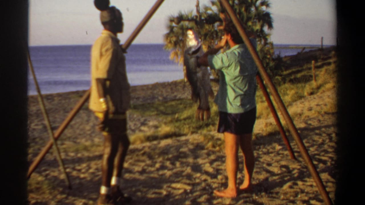 Two adult men stand on a sandy beach near a wooden tripod and hanging fish.