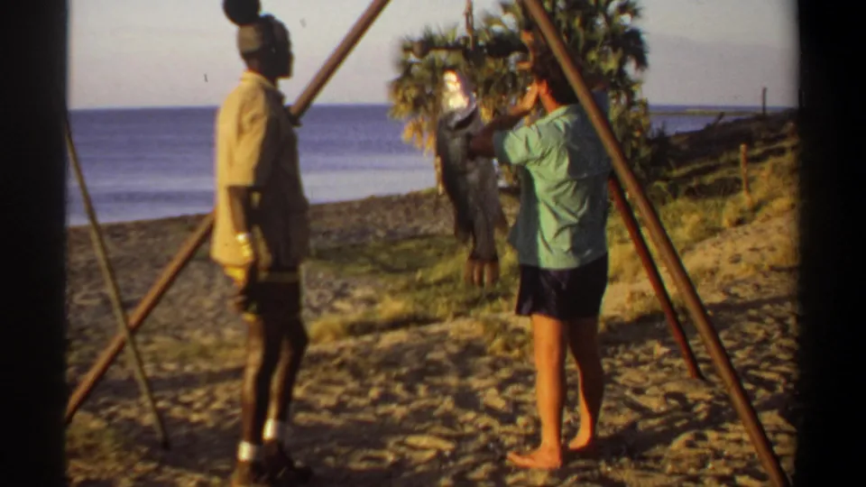 1969 Sudan-associated beach scene with two adult men beside a wooden tripod scale and a hanging fish.