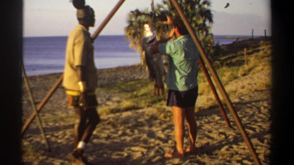 1969 Sudan-associated 8mm frame showing one man handling a fish on a wooden scale while another observes on a beach.