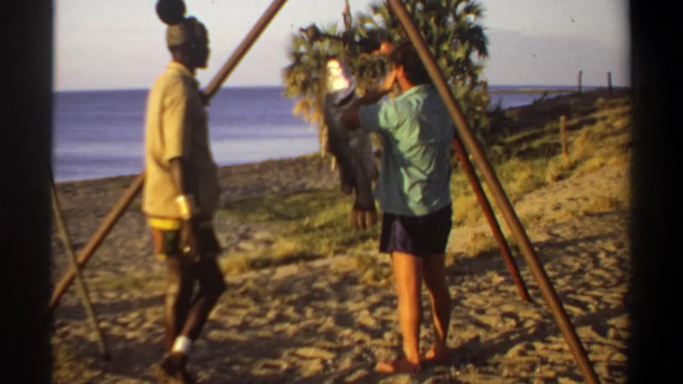 1969 Sudan-associated home-movie frame of two men beside a hanging fish on a makeshift beach weighing setup.