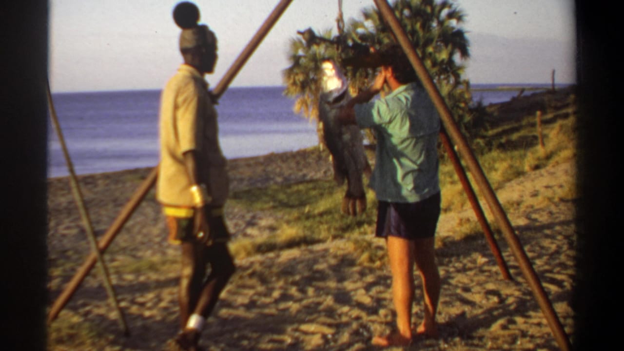 1969 Sudan home-movie frame showing two men beside a large hanging fish, with open water, sandy ground, and trees in the background.