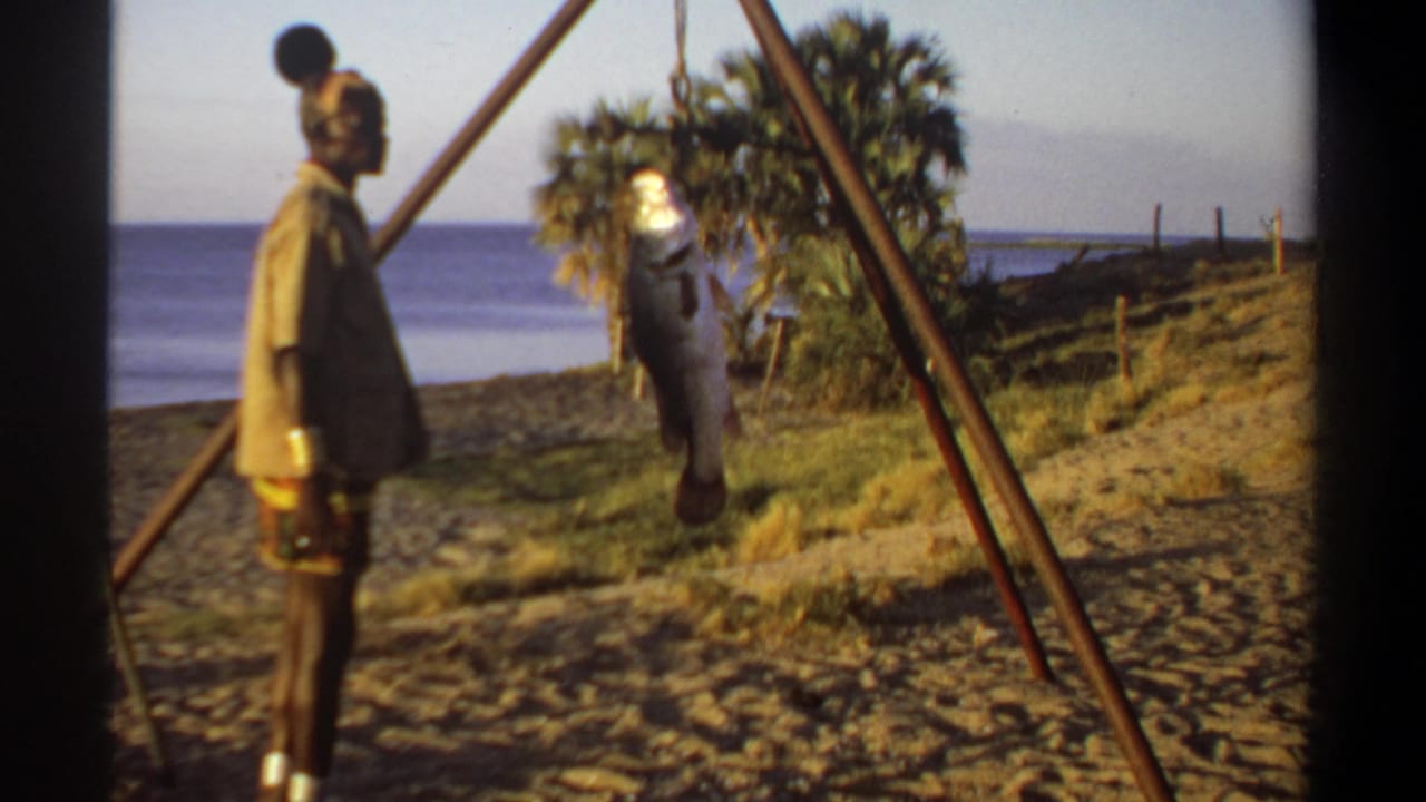 A man stands beside a large fish hanging from a wooden frame near water and trees.