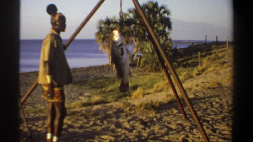1969 Sudan home-movie frame showing a man in distinctive headwear near a large hanging fish, with water and trees in the background.
