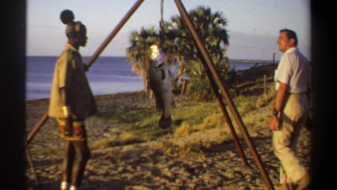 1969 Sudan-associated 8mm frame showing two adult men near a suspended large fish, with water and trees behind them.