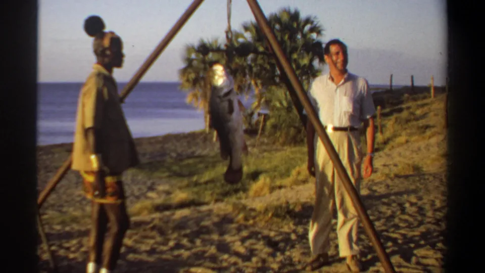 1969 Sudan-associated 8mm frame showing an adult man in light clothes near a hanging fish, with another man and water in the background.