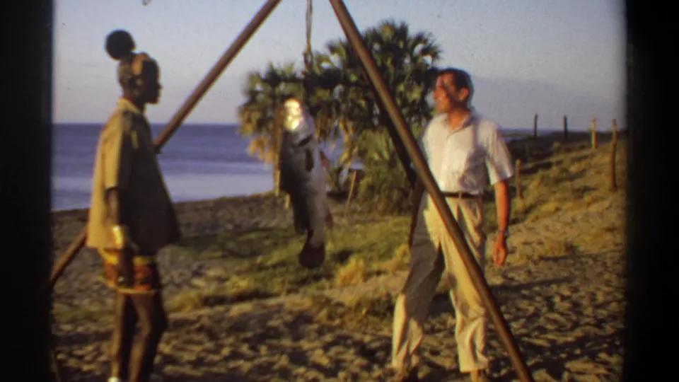 1969 Sudan-associated home-movie frame of two adult men near a tripod-suspended fish, with sandy ground, water, and trees visible.
