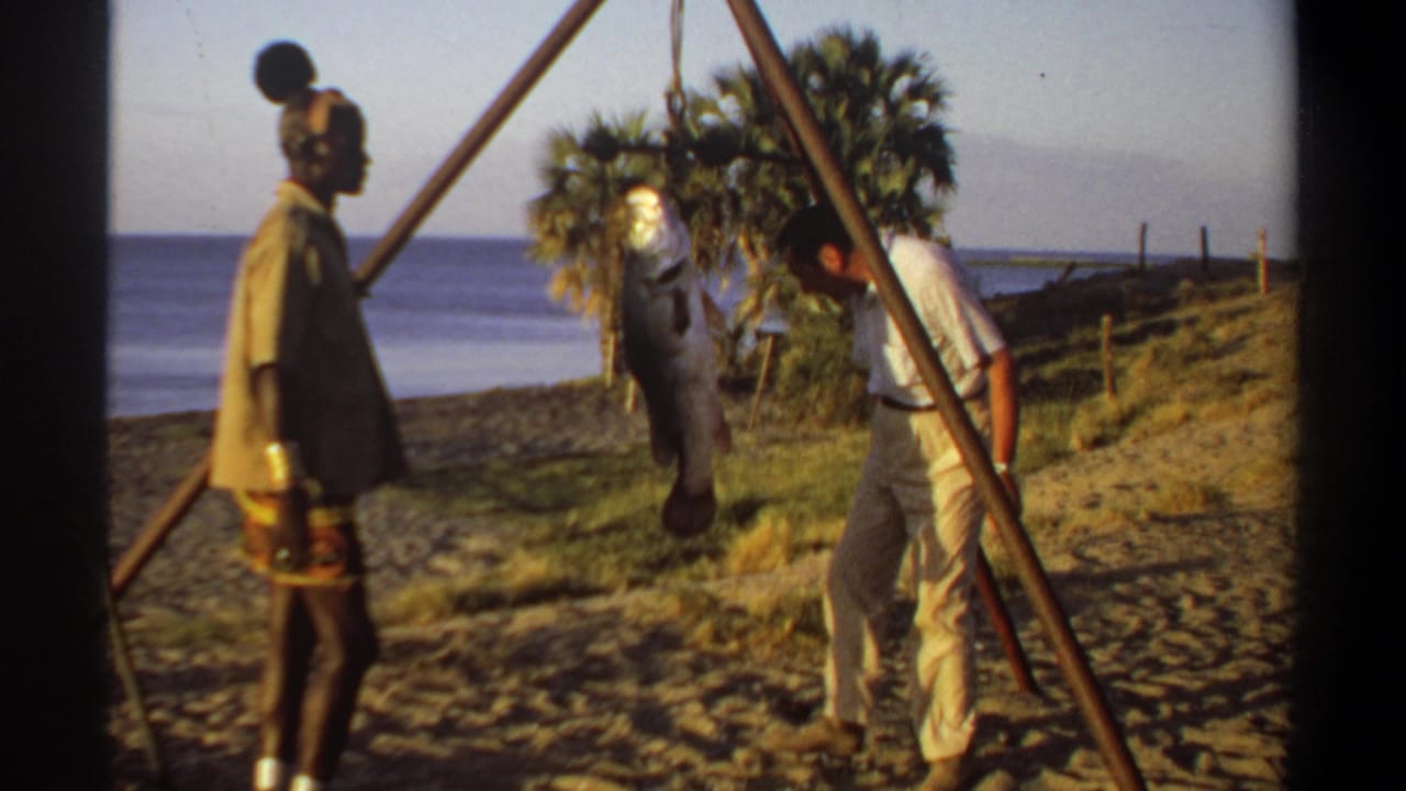 1969 Khartoum home-movie frame showing two men by a suspended large fish on a sandy riverside with palms and water behind them.
