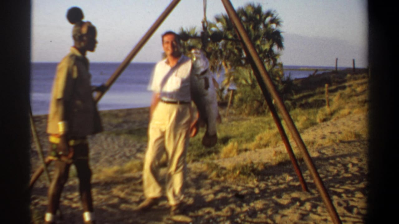 A man faces the camera beside a suspended large fish and another man near the tripod.