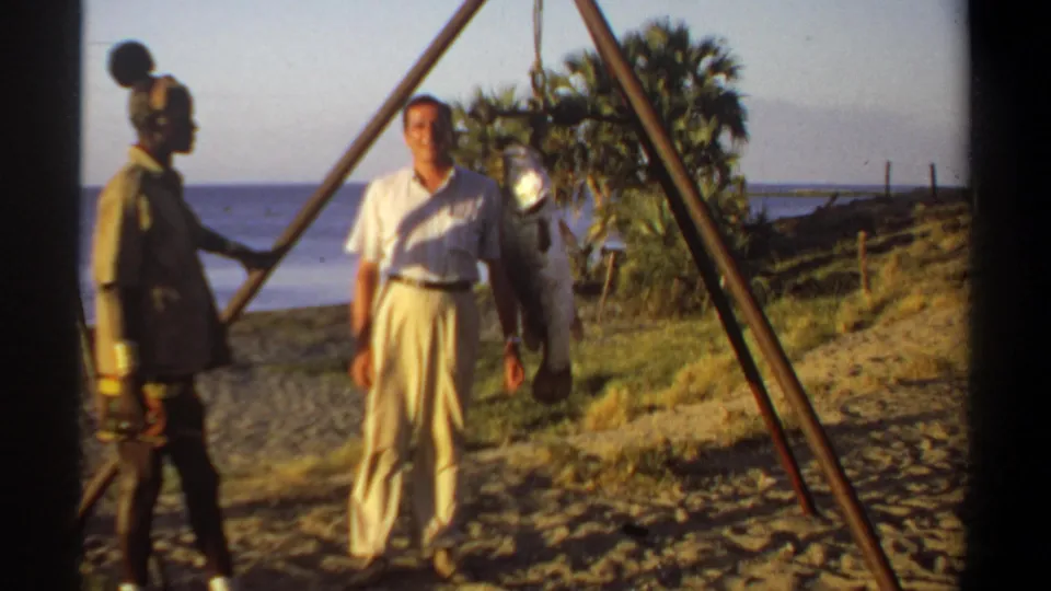 1969 Khartoum footage shows two men near a large fish suspended from a tripod, with river water and palm trees in the background.