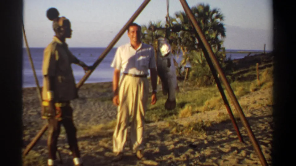 A late-1960s Khartoum home-movie frame shows a man standing beside a large suspended fish on a sandy riverside with palms behind.